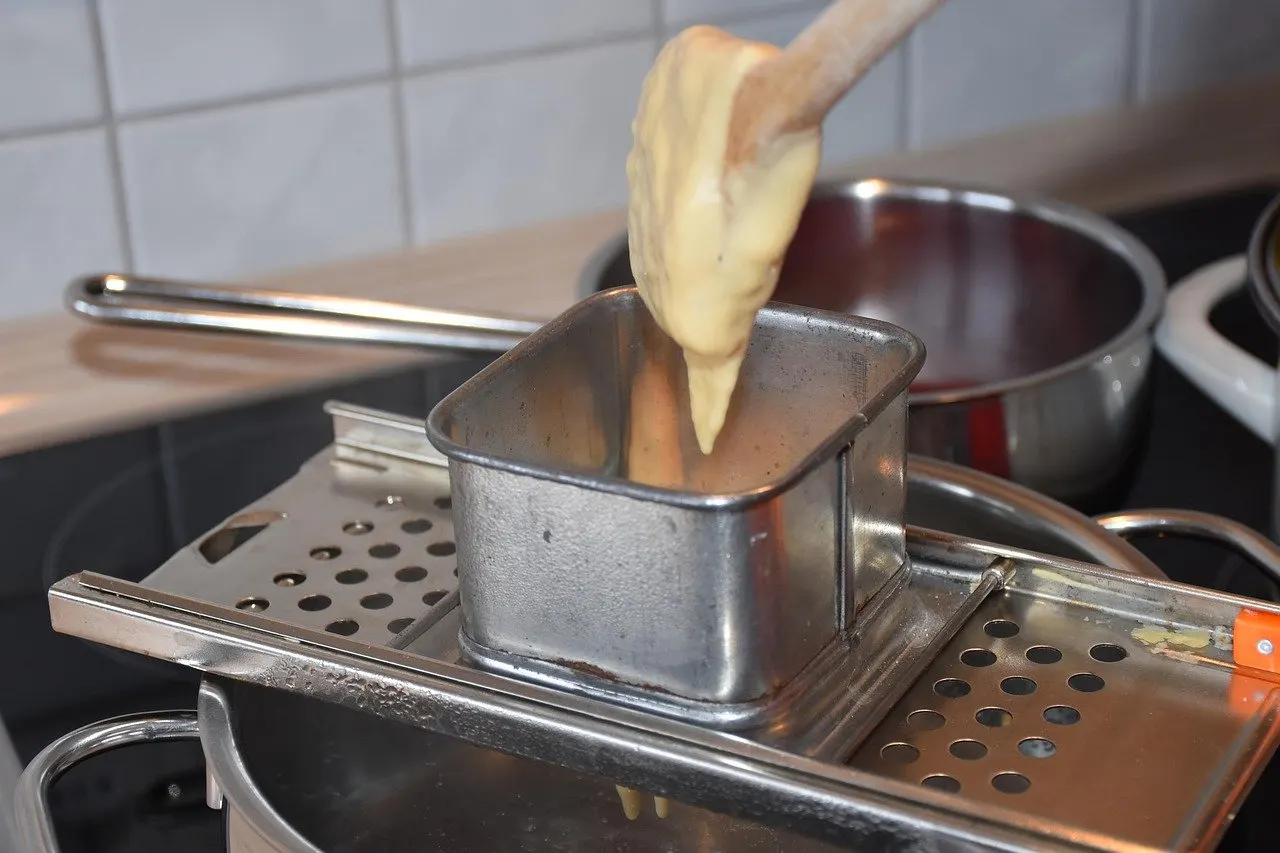 Zähflüssiger Spätzleteig läuft von einem Holzlöffel in den Schlitten eines Spätzlehobels, der auf einem Kochtopf liegt