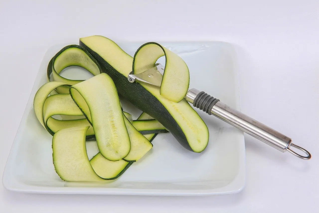 Halbe grüne Zucchini mit aufgelegtem Sparschäler und dünnen, abgezogenen Zucchinibändern auf einem eckigen weißen Teller